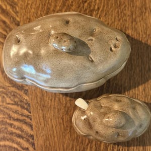 May include: A ceramic lidded container shaped like a potato with a speckled tan and brown glaze, featuring a small knob on the lid. Beside it sits a smaller matching ceramic lidded container with a tiny white spoon protruding from the side.