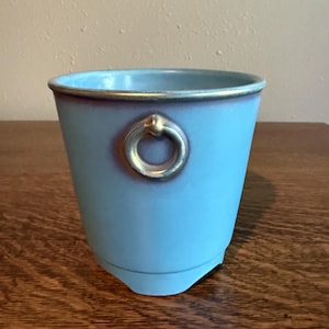 Peut inclure: Un pot de fleurs en céramique bleu clair avec une finition lisse et un dégradé violet subtil près du bord. Le récipient présente une poignée en forme d'anneau doré métallique fixée sur le côté et une bordure dorée assortie sur le dessus.