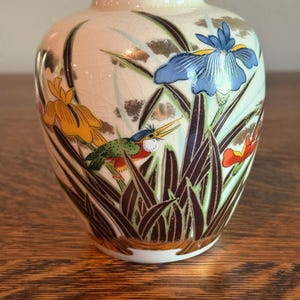 Può includere: Un piccolo vaso in ceramica color crema di forma rotonda con finitura smaltata a craquelé. La superficie presenta una scena dipinta a colori con un martin pescatore, fiori di iris blu e gialli e fogliame verde scuro con accenti dorati.