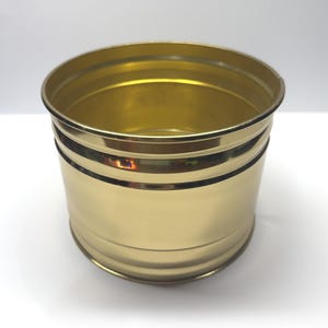 Puede incluir: Un recipiente cilíndrico de metal con un acabado dorado pulido. El objeto presenta una superficie lisa y reflectante con dos anillos horizontales en relieve que rodean la parte superior de la pared exterior y un borde superior abierto.
