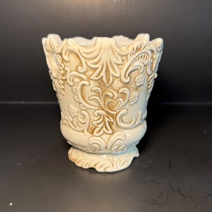 Puede incluir: Un recipiente de cerámica color crema con un intrincado patrón en relieve de hojas de acanto y motivos florales pequeños. El diseño presenta un borde festoneado y una base de pedestal, con tonos marrones que resaltan la textura.