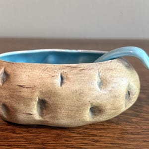 May include: A small, oval-shaped ceramic bowl with a textured, tan, potato-like exterior featuring small indentations. The interior of the bowl is glazed in a smooth, bright turquoise blue, and a matching turquoise ceramic spoon rests inside.