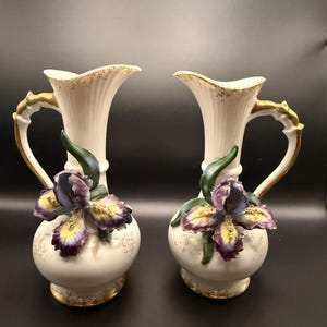 May include: A pair of cream-colored ceramic pitchers featuring ribbed necks, gold-painted handles, and rims. Each pitcher is adorned with a three-dimensional, hand-painted iris flower in shades of purple, yellow, and green, with gold accents.