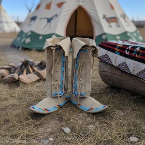 May include: A pair of tall, beige suede boots featuring light blue beaded panels with orange and black geometric patterns along the front, leather lace-up closures, and folded cuffs at the top, with additional beadwork along the base edges.
