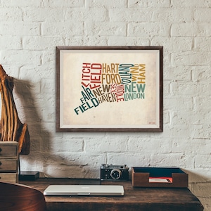 May include: A framed print of the state of Connecticut, filled with the names of cities in various colors. The print is in a dark brown frame and hangs on a white brick wall. A wooden desk with a laptop, camera, and desk organizer is in the foreground.