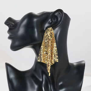 May include: A gold-colored, dangling earring with a mesh design. The earring is displayed on a black mannequin head. The mesh is made of small, connected circular pieces that create a flowing, draped effect. The earring has a rectangular top and a pointed bottom.