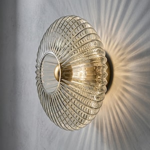 May include: A circular wall light featuring a ribbed, translucent glass shade with a warm amber tint. The centre of the fixture contains a smooth, polished gold-toned metal sphere, and the light emits a radial pattern of illumination.