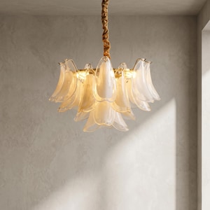Può includere: Un lampadario dal design floreale. L'apparecchio di illuminazione presenta più paralumi in vetro a forma di petalo in un morbido colore bianco crema, disposti attorno a una sorgente luminosa centrale. La montatura è dorata e l'apparecchio è sospeso a un cavo marrone.