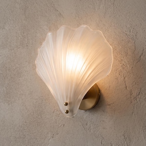 Puede incluir: Lámpara de pared con forma de concha. La pantalla de vidrio esmerilado presenta un diseño acanalado, emitiendo una luz cálida. Montada sobre una base circular de color latón, asegurada con dos pequeños tornillos dorados.