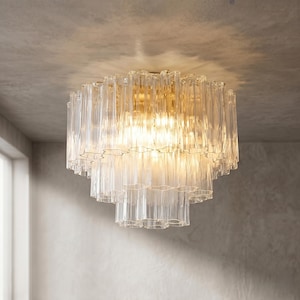 Könnte beinhalten: Ein mehrstufiger Kronleuchter aus transparenten Glasstäben mit einem goldfarbenen Metallfuß. Die Leuchte besteht aus zahlreichen vertikalen Glasstäben, die einen geschichteten, strukturierten Effekt erzeugen. Die Lichtquelle ist sichtbar und erzeugt ein warmes Leuchten.