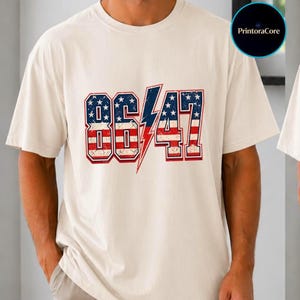 以下が含まれることがあります： クリーム色のTシャツ。前面に「86/47」の数字がヴィンテージ風のデザインでプリントされています。数字は星とストライプのパターンで埋められ、中央には赤い稲妻が描かれています。このシャツは、クラシックなクルーネックです。