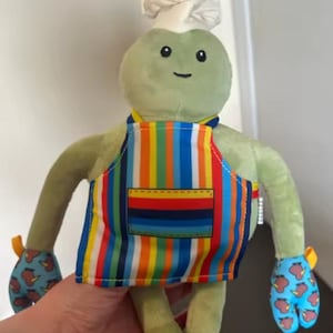 Puede incluir: Un peluche con forma de criatura verde que lleva un gorro de chef blanco, un delantal de rayas verticales coloridas con un bolsillo frontal y manoplas de cocina azules decoradas con un patrón repetitivo de pequeños kiwis marrones.