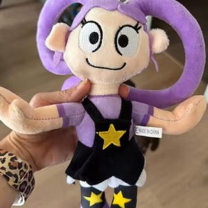 Op de afbeelding: Een pluche pop met paars haar in grote lussen, een lichte huidskleur en geborduurde gelaatstrekken. De pop draagt een paars shirt, een zwarte rok met een geel sterembleem en paarse laarzen met gele sterren als accenten.