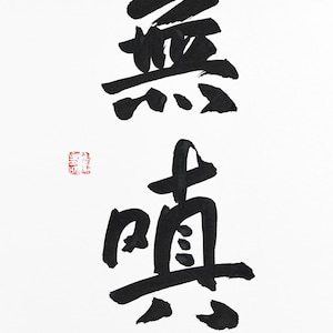 May include: A piece of white paper featuring black ink calligraphy of the Japanese kanji characters for 'mu' and 'shin'. To the left of the characters, there is a small, square red stamp with intricate, illegible white markings inside it.