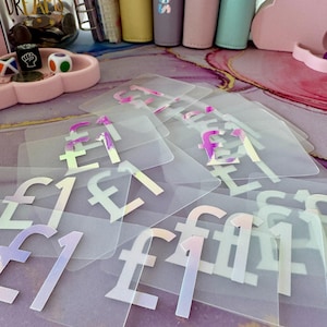 May include: A collection of clear plastic budget envelopes, each featuring an iridescent £1 symbol. The envelopes are arranged on a patterned surface. In the background, there are colorful binders and a jar with text.
