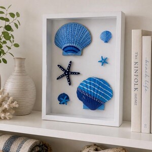 May include: A white rectangular shadow box frame containing a collection of blue and white painted seashells and starfish arranged on a white background. The display features two large fan-shaped shells, two small starfish, and one small round shell.