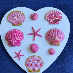May include: A white heart-shaped plaque decorated with various pink seashells and two pink starfish. The shells feature hand-painted designs including white dots, stripes, small flowers, and red hearts, with some edges accented in metallic gold paint.