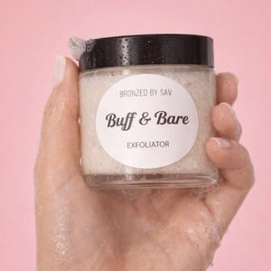 Puede incluir: Un tarro de vidrio transparente con tapa negra, sostenido por una mano. El tarro contiene una sustancia granulada blanquecina. La etiqueta dice "Bronzed by Sav", "Buff & Bare" y "Exfoliator". El fondo es rosa.