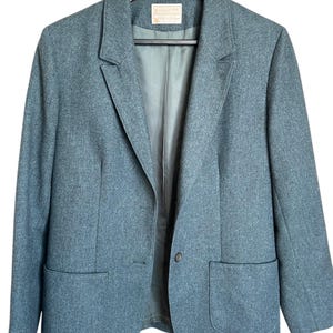 Puede incluir: Un blazer color verde azulado con dos bolsillos delanteros y cierre de un botón, colgado de una percha de madera. El blazer tiene un diseño de solapa clásico y una etiqueta dentro del cuello.