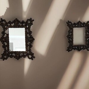 May include: Two rectangular wall mirrors featuring dark brown, ornate, scrollwork frames with intricate relief detailing. The mirrors are mounted side-by-side, with the left mirror being larger than the right, reflecting light on their surfaces.