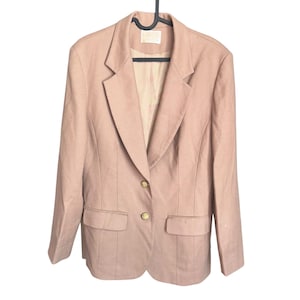 May include: A light pink blazer with a notched lapel, two front pockets, and gold-colored buttons. The blazer is hanging on a black hanger against a white background. The blazer has a tailored fit and a classic design.