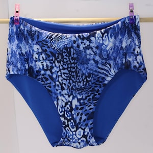 May include: High-waisted bikini bottoms featuring a vibrant blue and white animal print pattern. The garment is constructed with a solid blue interior lining and is displayed hanging from a wooden rod with two purple clips at the top corners.