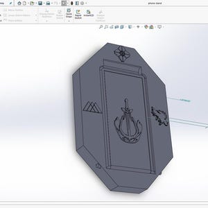 May include: A digital 3D CAD model of a rectangular phone stand displayed in SolidWorks software. The grey object features a central recessed panel with an engraved sword and harp emblem, flanked by a geometric logo and a bird-like crest design.