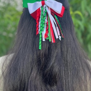 May include: A hair bow featuring a layered design in green, white, and red grosgrain ribbon. The accessory includes multiple hanging ribbon streamers in matching colors, with some ribbons featuring a spiral curl and others adorned with small beads.