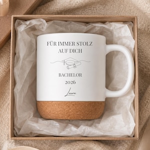 Op de afbeelding: Witte keramische mok met een kurken basis, met de tekst "FÜR IMMER STOLZ AUF DICH" en "Bachelor 2026" in het zwart. De mok wordt gepresenteerd in een geschenkdoos met een lint, ideaal als afstudeercadeau.
