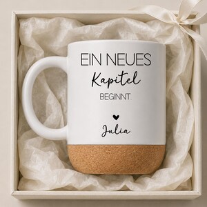 Peut inclure: Tasse en céramique blanche avec une base en liège, présentée dans un coffret cadeau avec un ruban. La tasse porte l'inscription "EIN NEUES Kapitel BEGINNT." et le nom "Julia" sous un symbole de cœur.