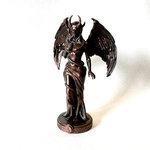 Può includere: Una statuetta color bronzo di una figura alata con corna, posizionata su una base circolare. La figura ha capelli lunghi, grandi ali piumate e un serpente avvolto attorno al corpo. La base presenta un piccolo emblema circolare con una mezzaluna.