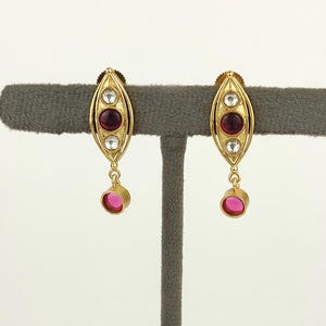 May include: A pair of gold-toned drop earrings featuring an elongated marquise-shaped frame. Each earring contains a central round dark red stone flanked by two clear white stones, with a single round dark red stone dangling from the bottom.
