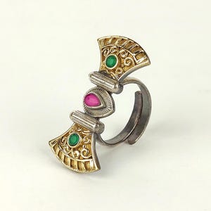 Puede incluir: Un anillo bicolor con una banda de color plateado y una pieza decorativa superior con detalles dorados en forma de abanico. El diseño incluye dos pequeñas piedras redondas verdes y una piedra central rosa en forma de lágrima en un marco de metal.