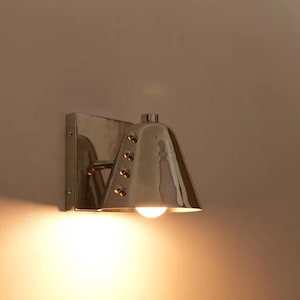 Peut inclure: Applique murale argentée polie avec un abat-jour en forme de cloche et une seule ampoule. L'applique est montée sur une plaque arrière carrée et comporte des rivets décoratifs. La lumière brille vers le bas, créant une lueur chaleureuse.