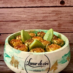 Op de afbeelding: Een decoratieve kom met een gemarmerd groen en wit exterieur, gevuld met goudkleurige, roosvormige items en groene bladaccenten. De kom heeft bladgoud accenten en de tekst "Lueur de cire - création" in een zwarte cirkel.