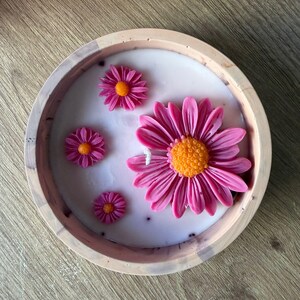 Peut inclure: Une bougie ronde dans un contenant clair présentant une surface de cire blanche lisse incrustée de quatre décorations en cire en forme de marguerites roses. La plus grande marguerite est à droite, trois plus petites sont à gauche.