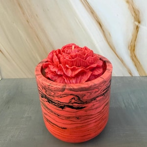 Peut inclure: Une bougie rouge en forme de pivoine est placée dans un récipient rond de couleur corail avec des marbrures noires. La bougie est d'un rouge vif et le récipient a une finition lisse et texturée. C'est un objet décoratif.