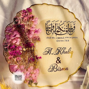 Op de afbeelding: Een ronde harsplaat met een geschulpte gouden rand, voorzien van ingebedde roze en paarse gedroogde bloemen aan de linkerkant. Het midden toont zwarte Arabische kalligrafie, de tekst "And We created you in pairs Quran 78/8" en "A. Khaliq & Bisma" in gouden schrift.