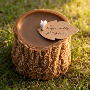May include: A brown candle shaped like a tree stump, with a smooth, light brown wax top. A small, leaf-shaped tag with the words "Especially for you" hangs from the wick. The candle sits on green grass.