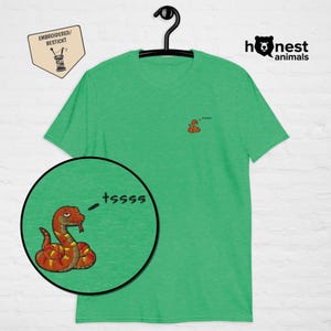 Pode incluir: Camiseta verde com um pequeno desenho de cobra bordada no peito e uma ilustração de cobra maior e detalhada em uma inserção circular. A cobra é laranja e amarela com a palavra "tsssss" ao lado. A camisa também tem o texto "Honest Animals".