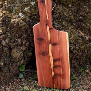 Puede incluir: Tabla de cortar de madera hecha a mano, de color marrón rojizo con una franja más clara en el centro. La tabla tiene un asa y se muestra contra un tronco de árbol, mostrando la veta natural de la madera. Mide unos 40 cm de largo.