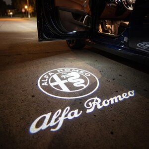 以下が含まれることがあります： 車のドアが開いており、白いアルファロメオのロゴが地面に投影されています。ロゴは「Alfa Romeo」の文字と、十字と蛇が描かれた円です。車は濃い青色です。