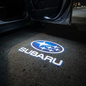 Puede incluir: Proyección en el suelo del logotipo blanco de Subaru, iluminado por la luz interior de un coche. El logotipo presenta un óvalo azul con estrellas blancas y el nombre de la marca en blanco. La puerta del coche está abierta.