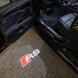 Puede incluir: Una puerta de coche abierta revela el interior y un logotipo proyectado en el suelo. El logotipo, "RS", es blanco con un contorno cuadrado rojo. El interior del coche presenta asientos y paneles de puerta de cuero negro.