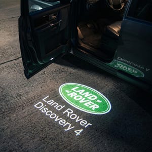 May include: A green and white Land Rover logo, with the words "Land Rover Discovery 4", is projected onto the ground. The image shows the open door of a dark-colored car, with the interior visible. The logo is illuminated.