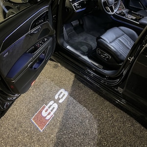 Puede incluir: Vista interior de un coche negro con la puerta abierta, que revela el logotipo "S3" iluminado proyectado en el suelo. El interior del coche cuenta con asientos de cuero negro y un salpicadero. El panel de la puerta es visible, junto con el umbral de la puerta.