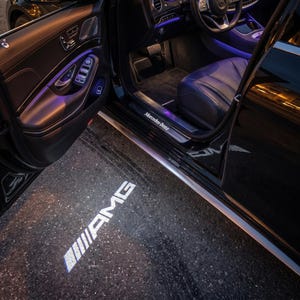 Könnte beinhalten: Eine offene schwarze Mercedes-Benz Autotür mit dem AMG-Logo, das in weißem Licht auf den Boden projiziert wird. Der Innenraum ist sichtbar, mit schwarzen Ledersitzen und blauer und violetter Ambientebeleuchtung.