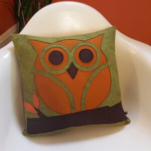 May include: A green and orange owl pillow with brown felt accents. The owl has large, round eyes and a branch with leaves.