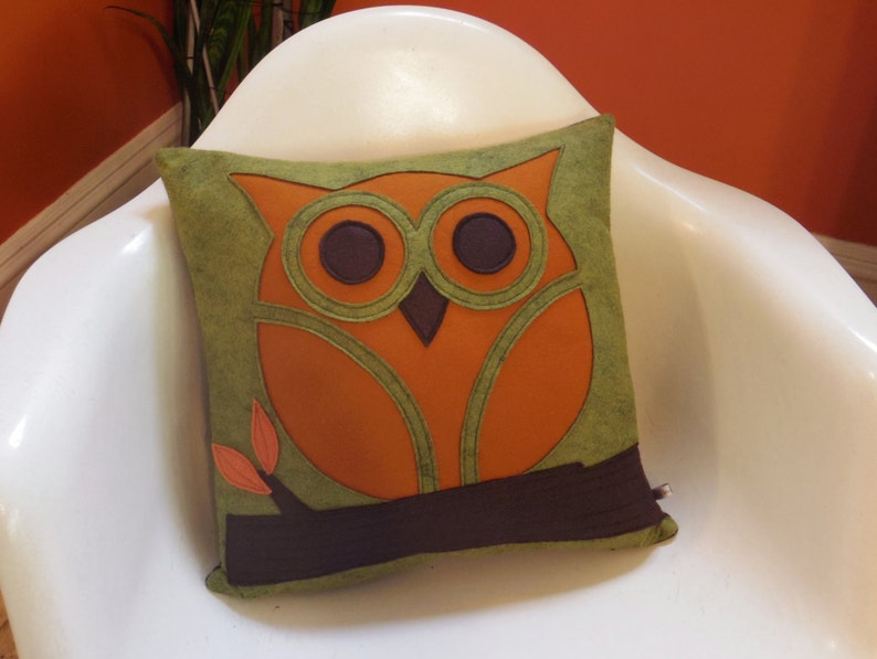 May include: A green and orange owl pillow with brown felt accents. The owl has large, round eyes and a branch with leaves.