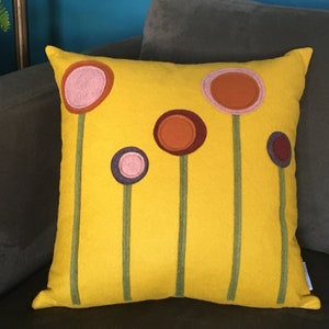 K&ouml;nnte beinhalten: Ein gelbes Kissen mit einem Design aus f&uuml;nf stilisierten Blumen in den Farben Rosa, Orange, Braun und Lila. Die Blumen sind gr&uuml;n umrandet.